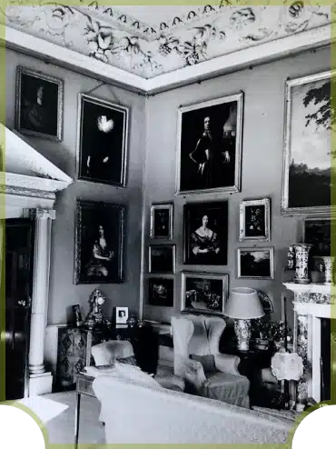 A black and white image of an elegant interior with framed paintings on the walls, ornate moldings on the ceiling, and classical furniture. An armchair, a lamp on a side table, and decorative vases add to the room's vintage charm.