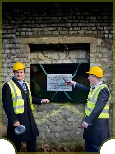 Two men in suits and high-visibility vests, wearing yellow construction helmets, unveil a plaque mounted on a stone wall. One holds a hard hat, and the other points at the plaque, marking an opening ceremony or dedication event.