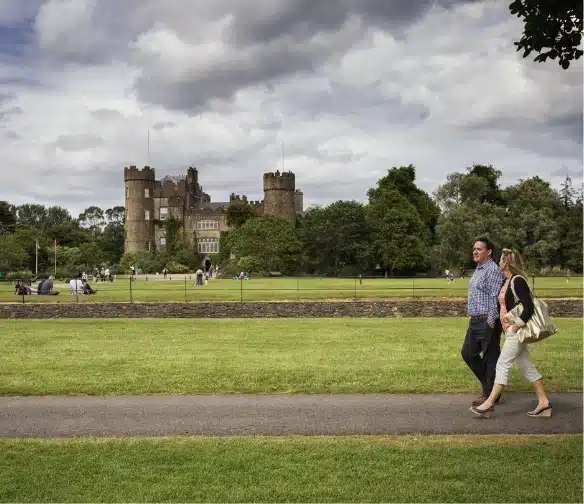 A couple strolls along a pathway in a park with lush green grass and trees. In the background, the majestic Malahide Castle stands proudly under a cloudy sky, with visitors exploring its historic grounds.