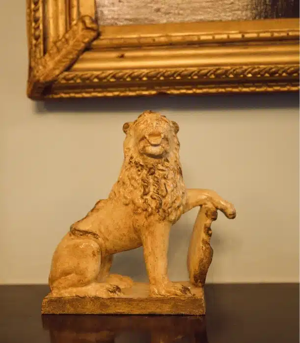 A small, intricately detailed lion sculpture sits on a dark surface, one paw raised over a shield. Behind it looms a section of a framed painting with a gold frame, reminiscent of the regal charm found within Malahide Castle.