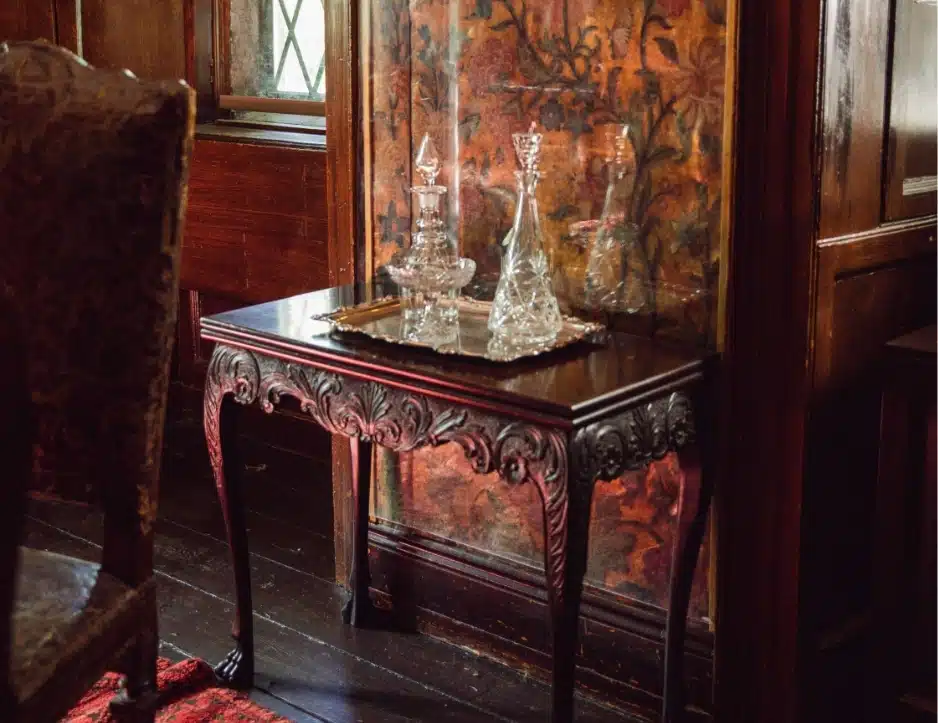 In a dimly lit room reminiscent of Malahide Castle, an ornate wooden table with carved legs holds a silver tray adorned with crystal decanters. Floral-patterned wallpaper surrounds the scene, while a carpet and an upholstered chair are partially visible in this elegant setting.