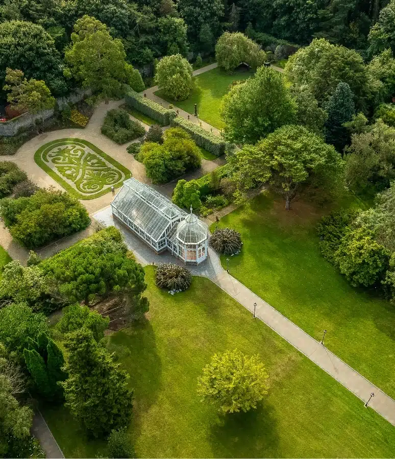 Aerial view of the lush garden at Malahide Castle, featuring a central glass conservatory, surrounded by vibrant green lawns and winding pathways. An ornate, symmetrical design is visible with manicured trees and neatly trimmed hedges, echoing the elegance of this historic estate.