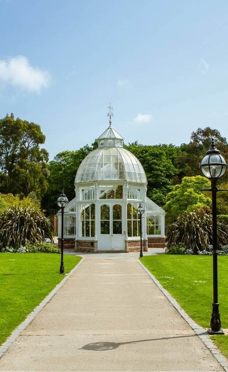 A white Victorian-style glasshouse stands at the end of a paved path at Malahide Castle, surrounded by manicured green grass and two vintage street lamps. Lush trees and plants frame the scene under a clear blue sky, echoing the serene charm of this historic estate.