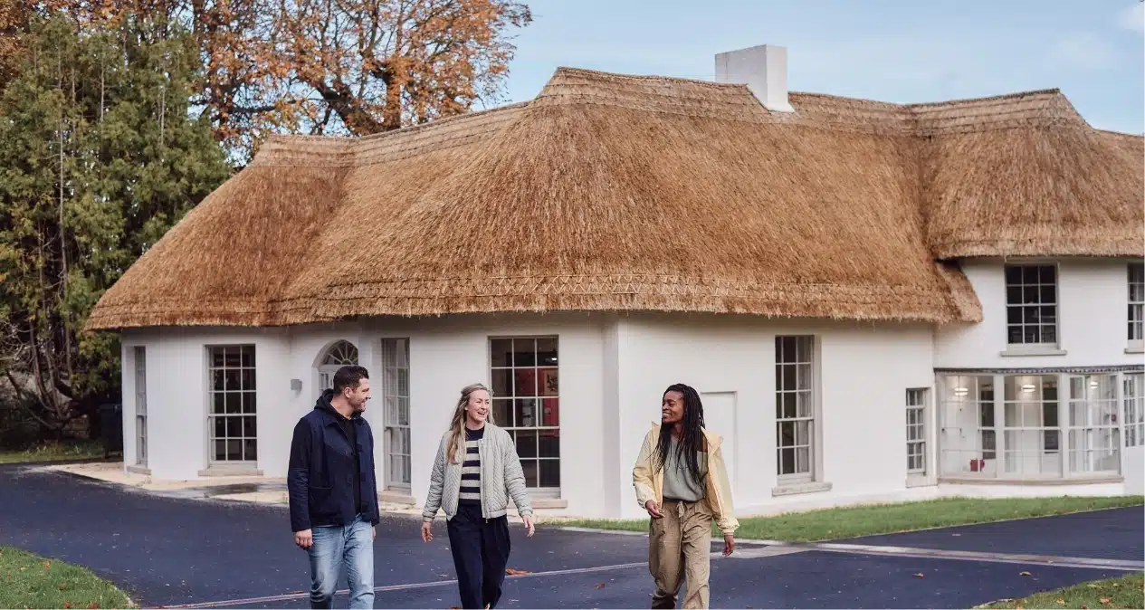 Three people walk and talk outside a white building with a thatched roof, reminiscent of the structures near Malahide Castle, all surrounded by lush trees and a vibrant blue sky.