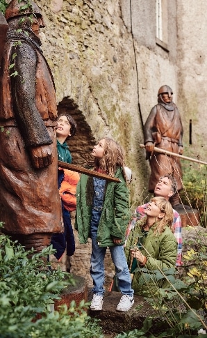 A family with two adults and two children observes large wooden statues outside Malahide Castle. The statues resemble historical figures in a rustic setting with stone walls and greenery. The children, wearing jackets, look up with curiosity.