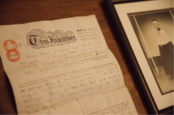 A vintage document titled "This Indenture" is laid out on a wooden table next to a framed black-and-white portrait of a man, evoking the storied charm of places like Malahide Castle. The handwritten text on the document appears to be an old legal agreement.