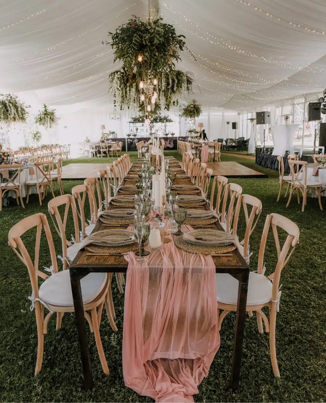 A beautifully decorated wedding reception at Malahide Castle features a long wooden table with a pink runner and elegant place settings under a tent. Wooden chairs surround the table, lush greenery with lights hangs from the ceiling, and the grass-covered floor completes the enchanting scene.