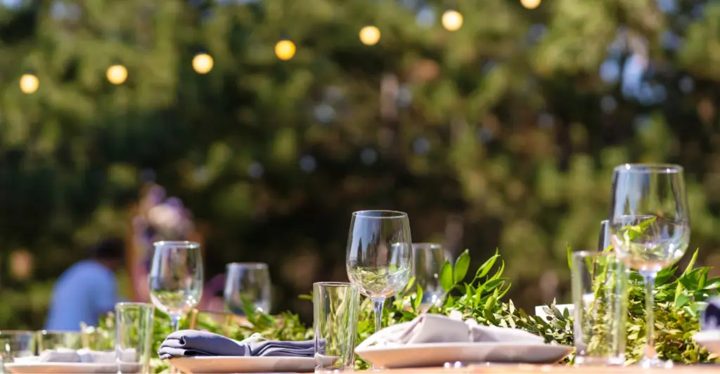 A beautifully set outdoor dining table at Malahide Castle features empty wine glasses, cloth napkins, and lush greenery as a centerpiece. Soft, glowing string lights hang in the background, creating a warm and inviting atmosphere.