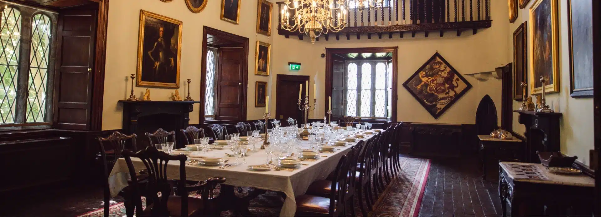 A grand dining room in Malahide Castle boasts a long, elegantly set table surrounded by wooden chairs. The room features a large chandelier, ornate paintings, tall diamond-patterned windows, and a high ceiling with wooden beams.