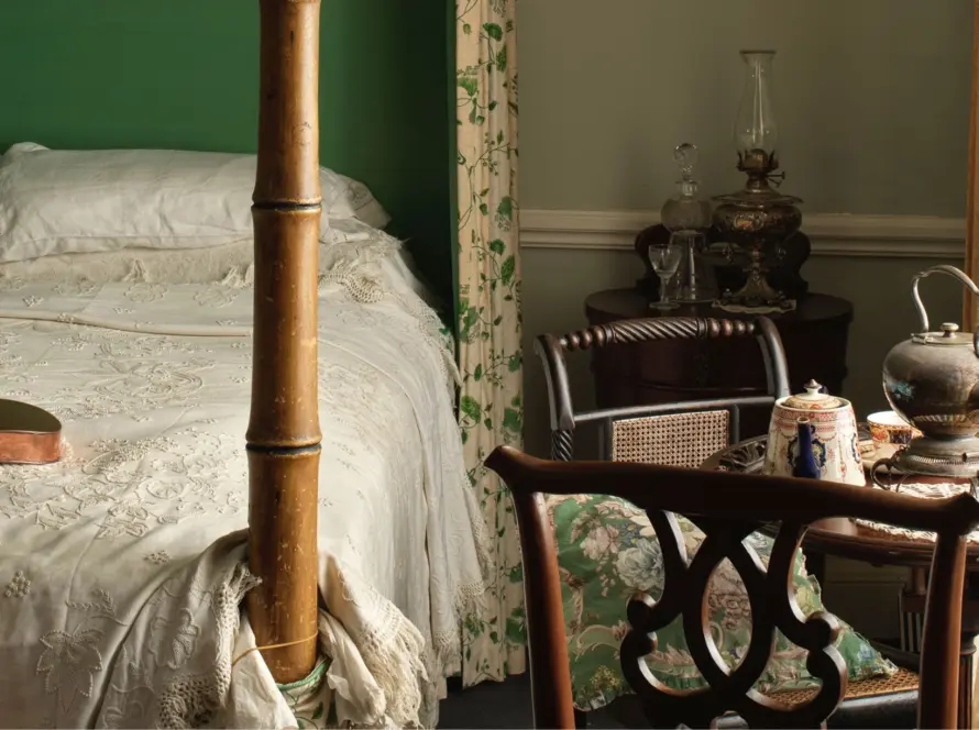 A vintage bedroom scene reminiscent of Malahide Castle, featuring a four-poster bed with white lace bedding and a copper warming pan. A small table holds a silver teapot, cups, and saucers. The room is styled with a green accent wall, floral curtains, and an antique chair nearby.