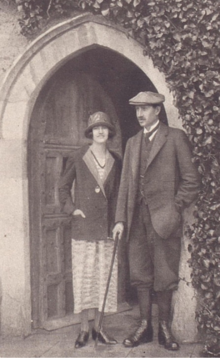 A sepia-toned image captures a man and woman standing before an ivy-clad, arched wooden door at Malahide Castle. The woman dons a long coat, hat, and skirt, while the man wears a suit with a flat cap, holding a walking stick.