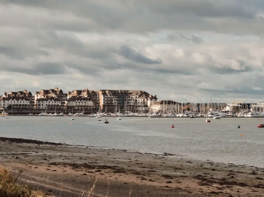 Scenic view of a coastal town near Malahide Castle, with several boats anchored in a calm harbor. The shoreline is lined with charming buildings under a cloudy sky, and the foreground features a beach with some vegetation.