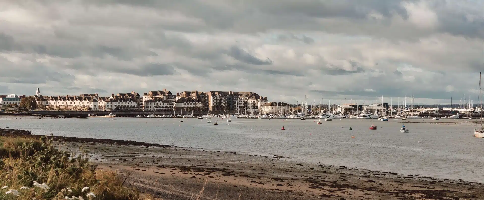 Scenic view of a coastal town near Malahide Castle, with several boats anchored in a calm harbor. The shoreline is lined with charming buildings under a cloudy sky, and the foreground features a beach with some vegetation.