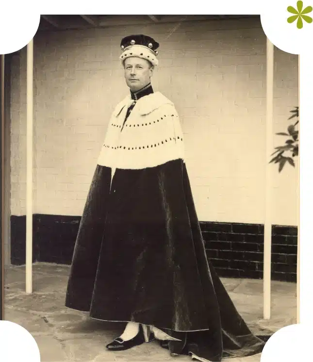 A person wearing a royal ceremonial robe and crown poses for a photo in front of the white-brick wall, reminiscent of Malahide Castle's historic charm. Their dark robe, adorned with white fur trim, adds to the regal ambiance against the setting's elegant black skirt at the bottom.