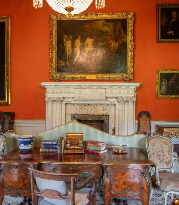 A richly decorated room in Malahide Castle features an ornate fireplace, two framed paintings on a red wall, a chandelier, and a wooden table with books and decorative items. Vintage chairs surround the table, perfectly complementing the classic decor.