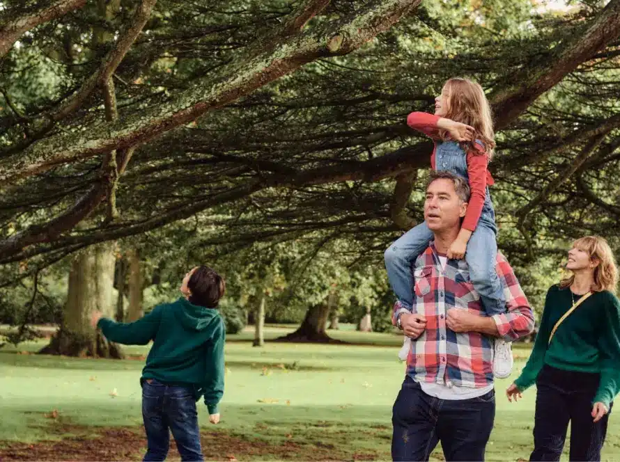 A family enjoys a day at Malahide Castle's park. A man carries a girl on his shoulders while another child looks up at a tree. A woman accompanies them, all surrounded by lush greenery.