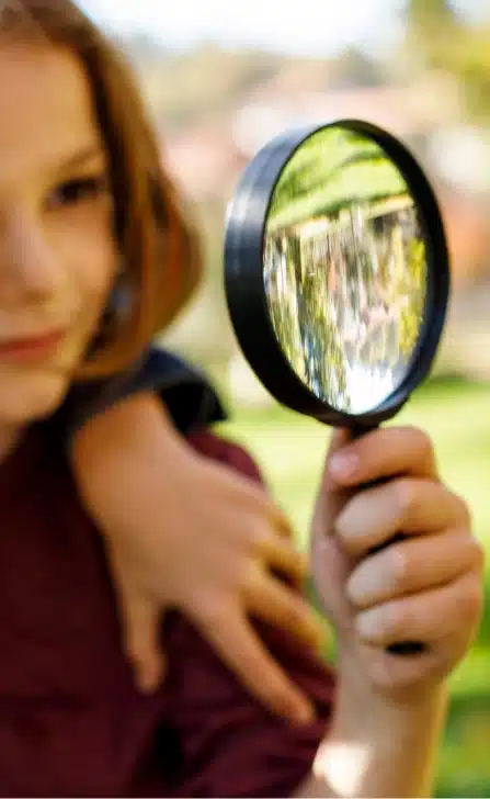 A child holds a magnifying glass, reflecting the lush greenery of Malahide Castle's gardens. Another child's arm is draped over the first one's shoulder in a tender gesture. The scene unfolds outdoors, with a softly blurred background of grass and towering trees whispering tales from the past.