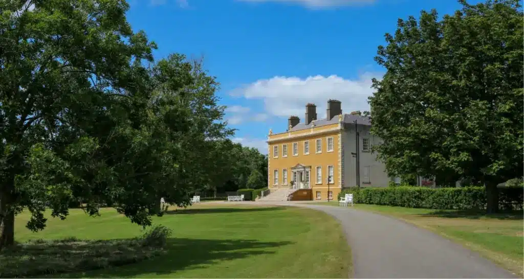 A large, historic yellow mansion reminiscent of Malahide Castle stands amidst a lush green lawn, with a paved path leading to its entrance. Tall trees border the scene under a bright blue sky with scattered clouds.