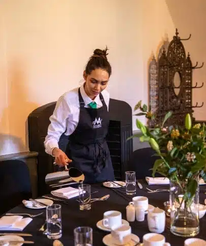 A waitress with a bun hairstyle, wearing a black apron, gracefully pours water into a glass at an elegantly set table with white cups and plates. The scene, reminiscent of Malahide Castle's charm, features a lovely floral arrangement and decorative wall elements in the background.