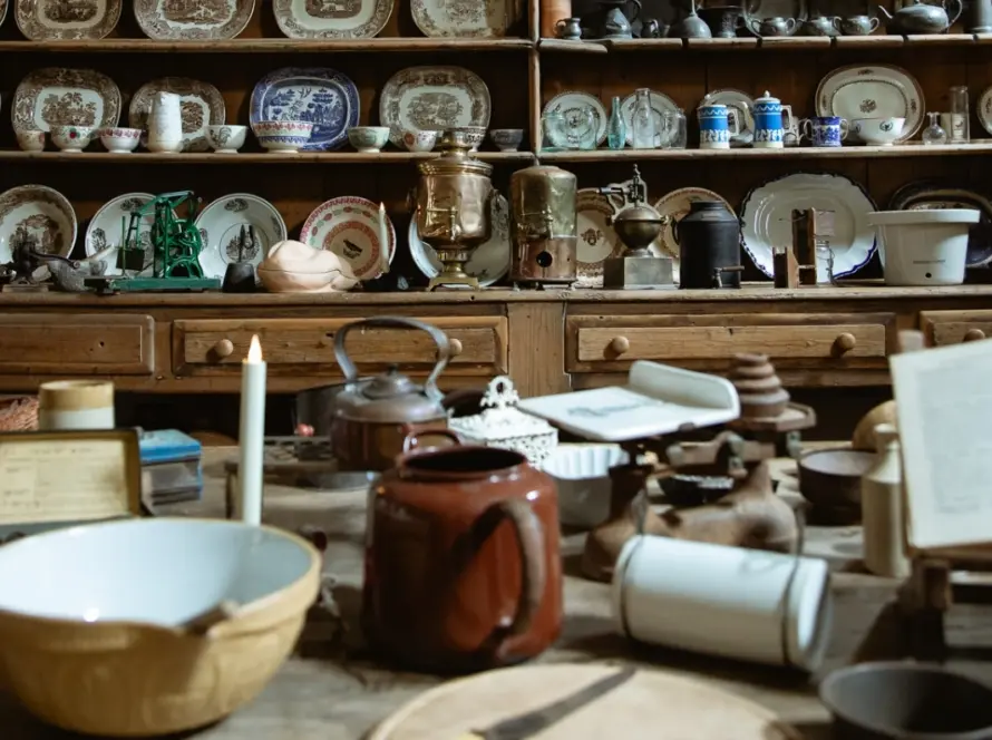 A cozy antique kitchen reminiscent of Malahide Castle, filled with vintage crockery, pots, and utensils. A wooden table displays an open book, teapot, bowls, and candlestick. Shelves in the background showcase patterned plates and miscellaneous kitchenware.