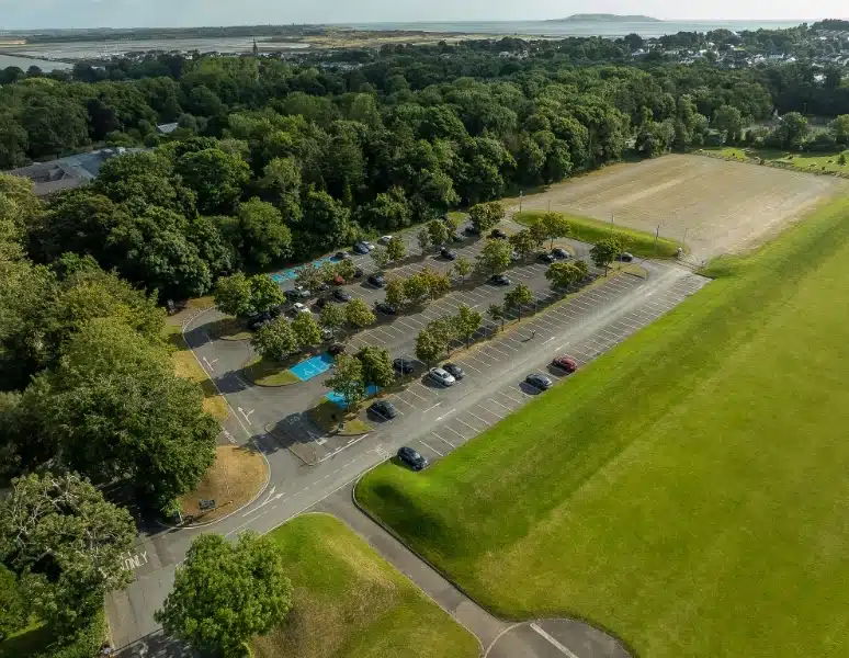 Aerial view of a parking lot adjacent to the sprawling green park area near Malahide Castle. Several cars are parked in the lot, including designated blue handicap spaces. The scene is surrounded by trees and the sky is clear, offering a serene backdrop for this historic site.