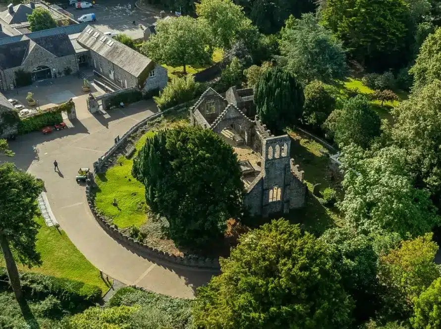 Aerial view of a park featuring historic stone ruins near Malahide Castle, surrounded by lush green trees. Nearby, modern buildings with sloped roofs are connected by winding pathways. The scene is peaceful, set under a clear sky.