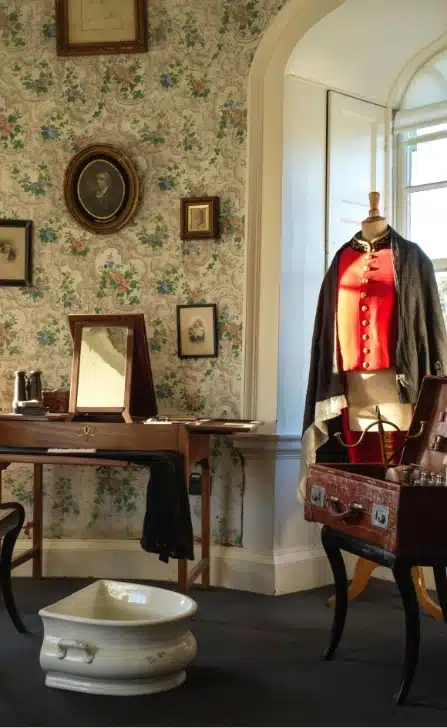 A vintage room reminiscent of Malahide Castle, with floral wallpaper, features a wooden vanity with a mirror, a red military-style coat on a mannequin, framed portraits, a suitcase, and a white ceramic basin on the floor. Sunlight streams through the window.