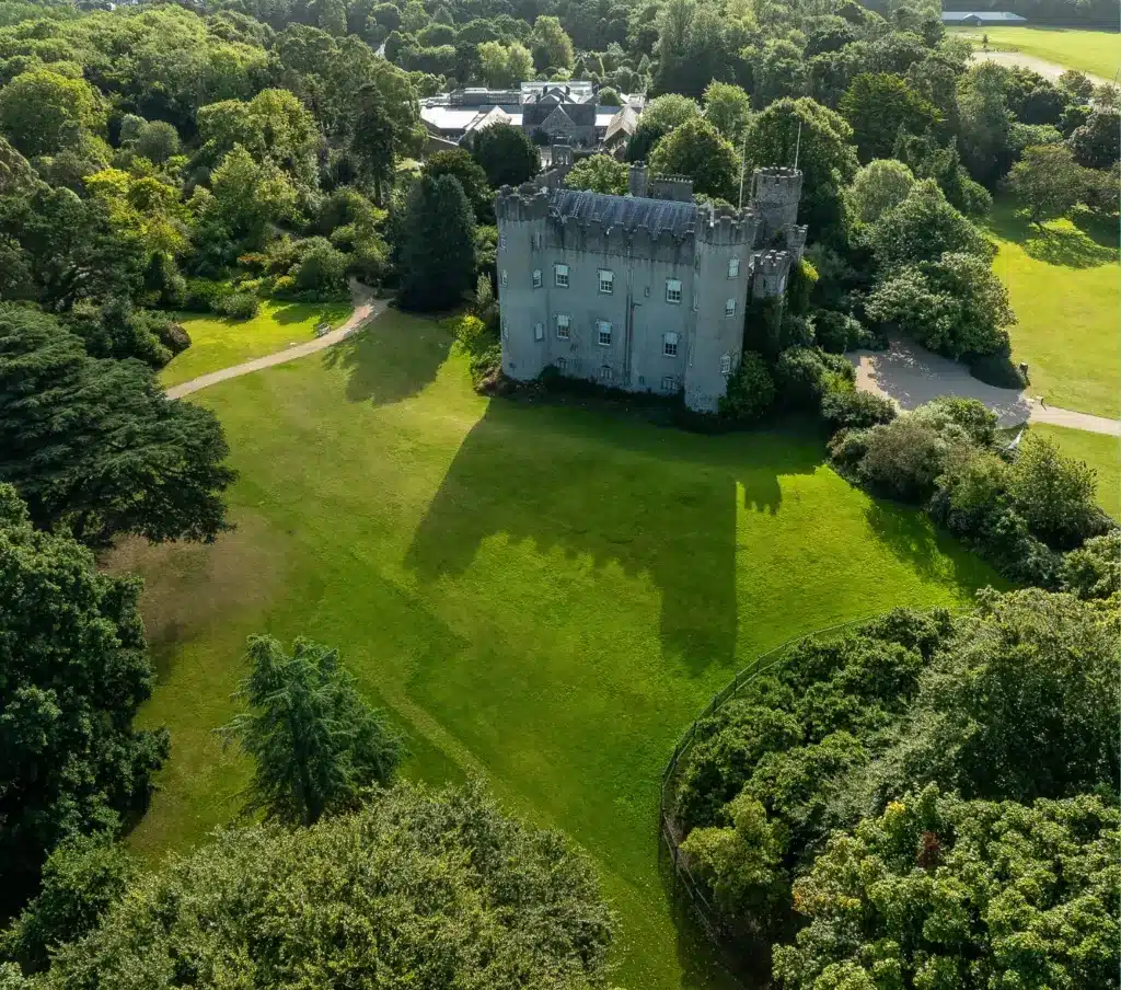 An aerial view of Malahide Castle reveals its medieval stone grandeur surrounded by lush green trees and manicured lawns. The castle casts a shadow on the grass, with paths meandering through the landscape, while dense forest envelops one side.