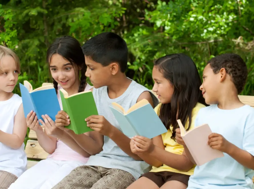 Five children are sitting on a wooden bench in the park near Malahide Castle, each engrossed in reading a book. They are surrounded by lush green foliage, and the weather appears sunny and pleasant.