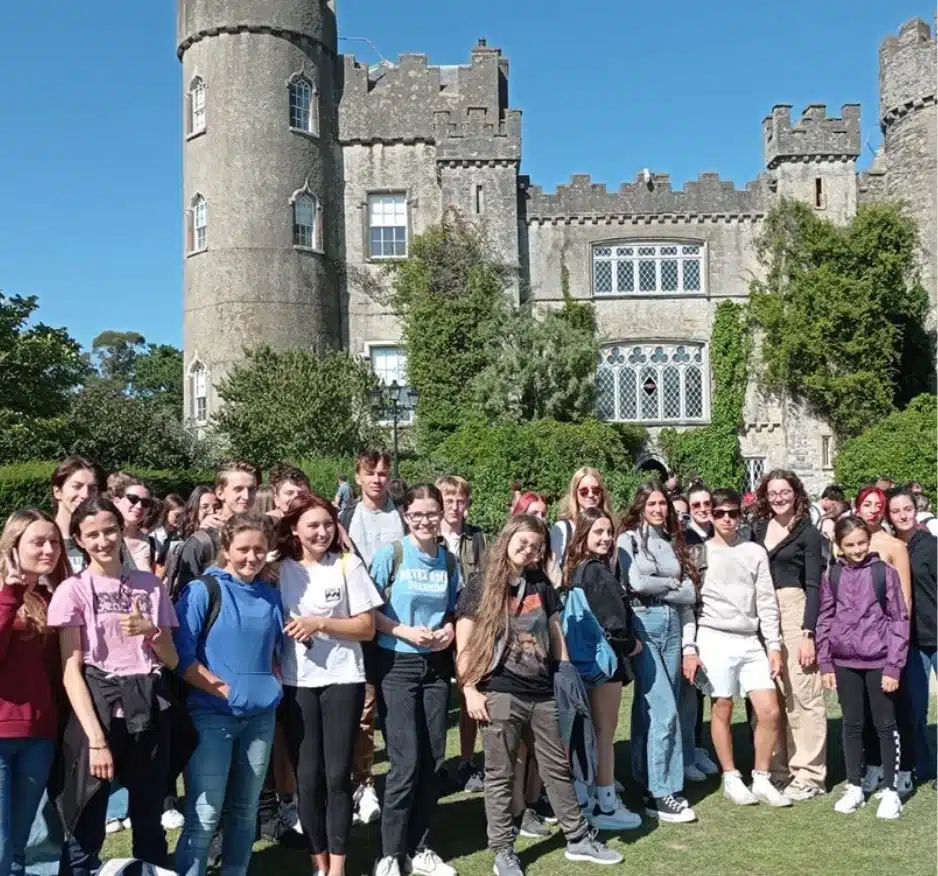 A large group of people stands in front of the historic Malahide Castle, with its elegant turrets and stone walls. On a sunny day, they smile warmly, dressed casually amidst the lush greenery that envelops this picturesque landmark.