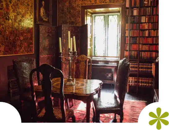 A quaint library in Malahide Castle features vintage wooden furniture, with a small table adorned by a candelabra, surrounded by chairs. Books fill shelves beside a stained glass window, while patterned wallpaper adds warmth to the room.