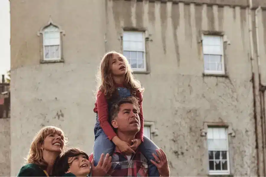 A family stands in front of Malahide Castle, its historic stone crenellations towering above. A man carries a young girl on his shoulders, while a woman and a boy stand nearby, looking up. Framed by trees, the scene is light and cheerful.