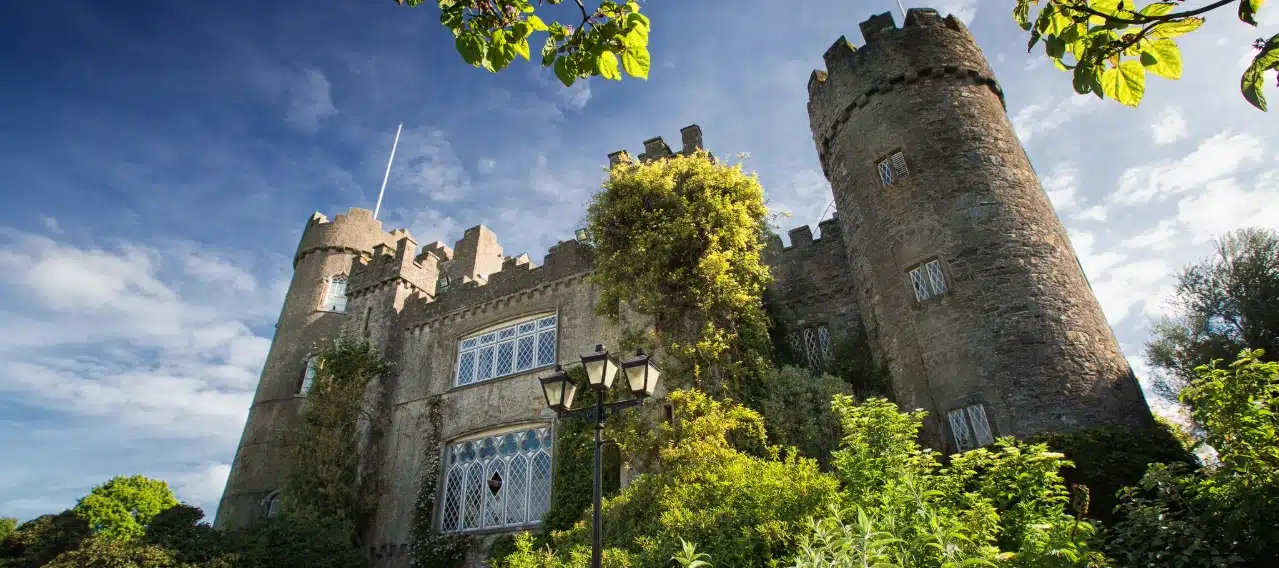 A majestic stone castle with cylindrical towers, reminiscent of Malahide Castle, stands proudly against a blue sky. Sunlight highlights its detailed architecture, overgrown with ivy and surrounded by lush greenery. A streetlamp is visible in the foreground.