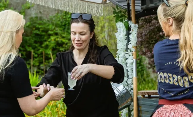 A group of women gathered outdoors near a mobile bar at Malahide Castle. One woman in black is serving drinks to another woman with blonde hair. The bar, adorned with white flowers, stands out against the lush greenery in the background.