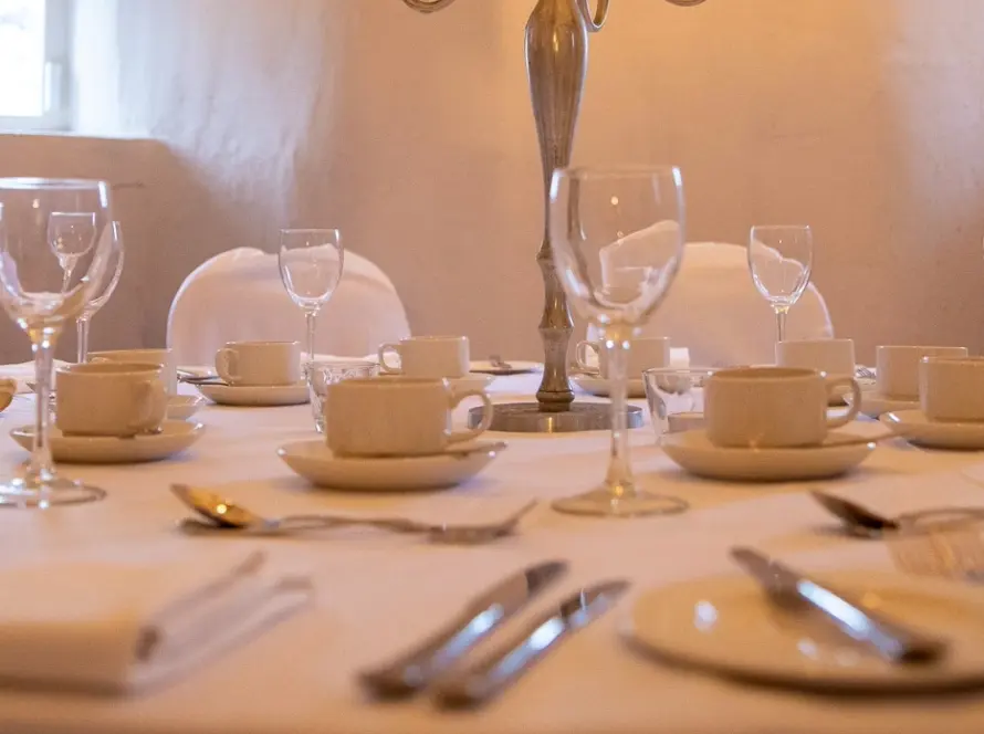 A round table, reminiscent of those in Malahide Castle, is elegantly set for a formal event. It features white tablecloth and napkins, fine china, glassware, and silverware. Plates, wine glasses, teacups, and saucers are arranged around the softly lit backdrop.