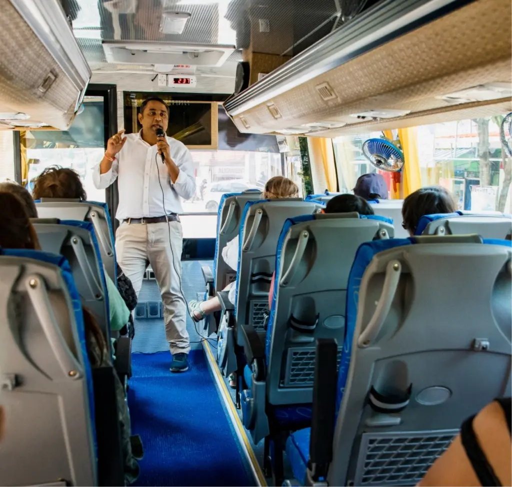 A man at the front of a tour bus, speaking into a microphone, highlights the upcoming visit to Malahide Castle. The bus interior is bright, with blue accents and overhead compartments. Passengers face forward, listening attentively to the historical insights being shared.