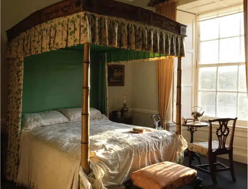 A sunlit castle room features a vintage four-poster bed draped in floral fabric and green curtains. The aging white bedding adds charm. The room includes a wooden chair, a small table, a stool, and a large window with sheer curtains that flood the space with natural light.