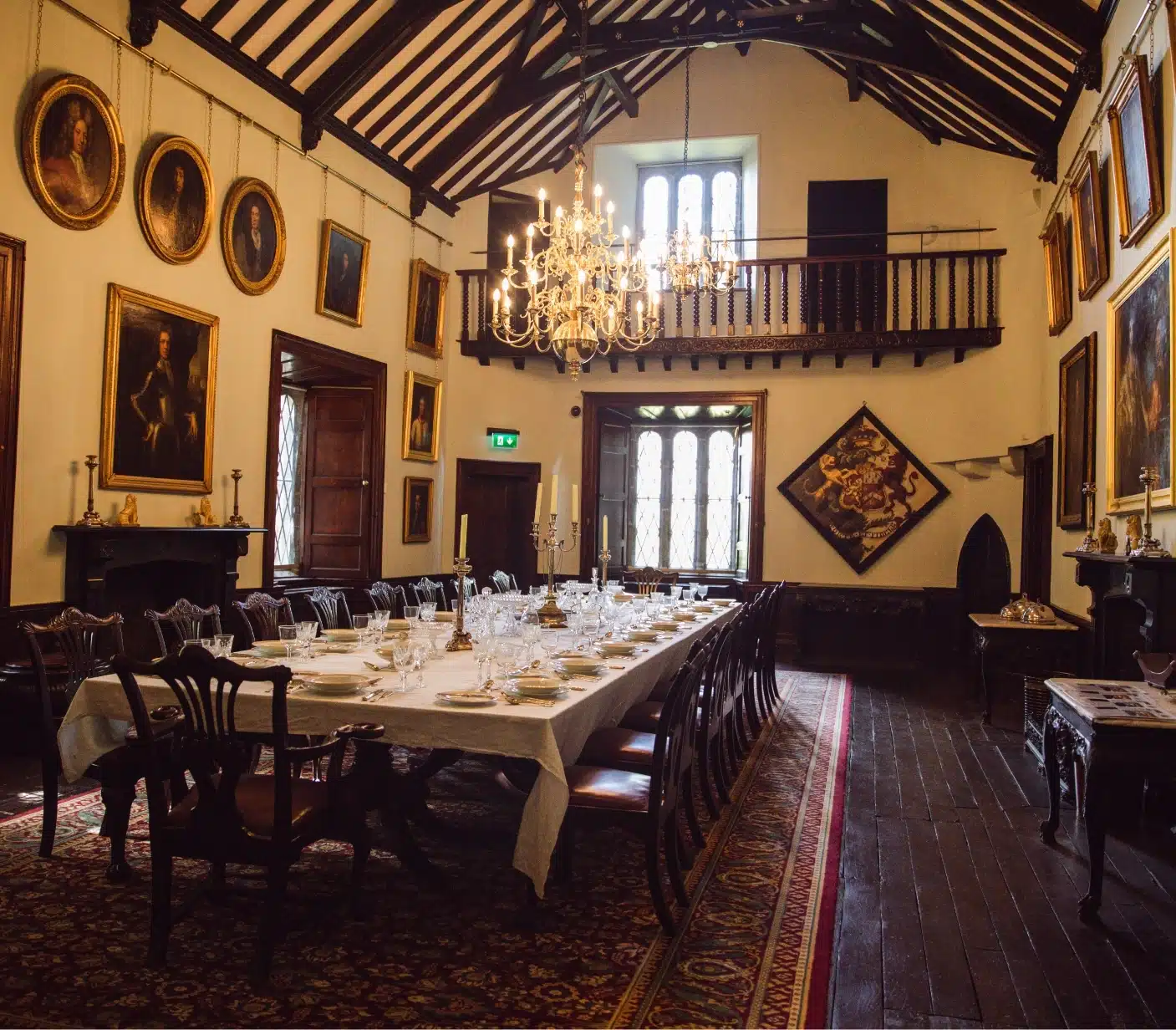 A grand dining hall with a long table set for a meal, surrounded by ornate wooden chairs. The walls are adorned with framed portraits. A large chandelier hangs from the high, beamed ceiling, and an upper balcony overlooks the room.