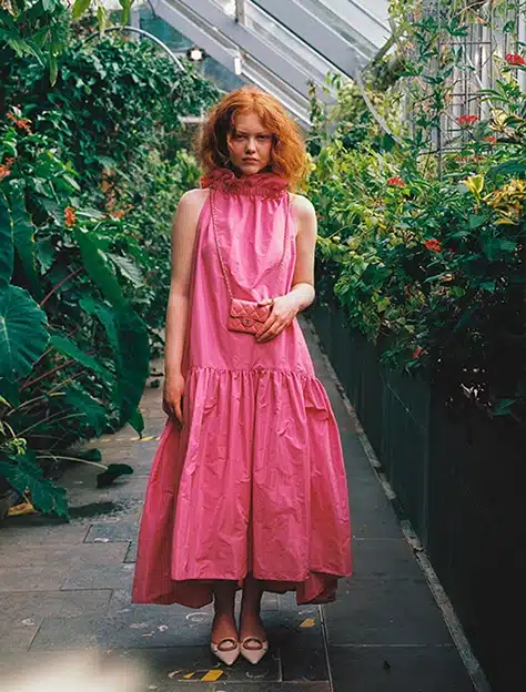 Amid the enchanting ambiance of Malahide Castle's greenhouse, a person with curly red hair stands elegantly in a long, bright pink dress, holding a small pink bag. Lush green plants and vibrant flowers line the path on both sides.