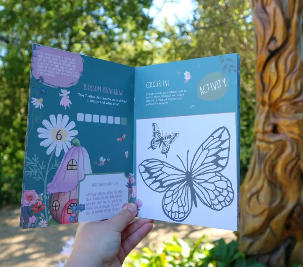 A hand holds an open activity booklet outdoors near Malahide Castle. The left page features a colorful illustration of a mushroom house and text with a flower pattern. The right page shows a butterfly drawing for coloring, with trees and sunlight visible in the background.