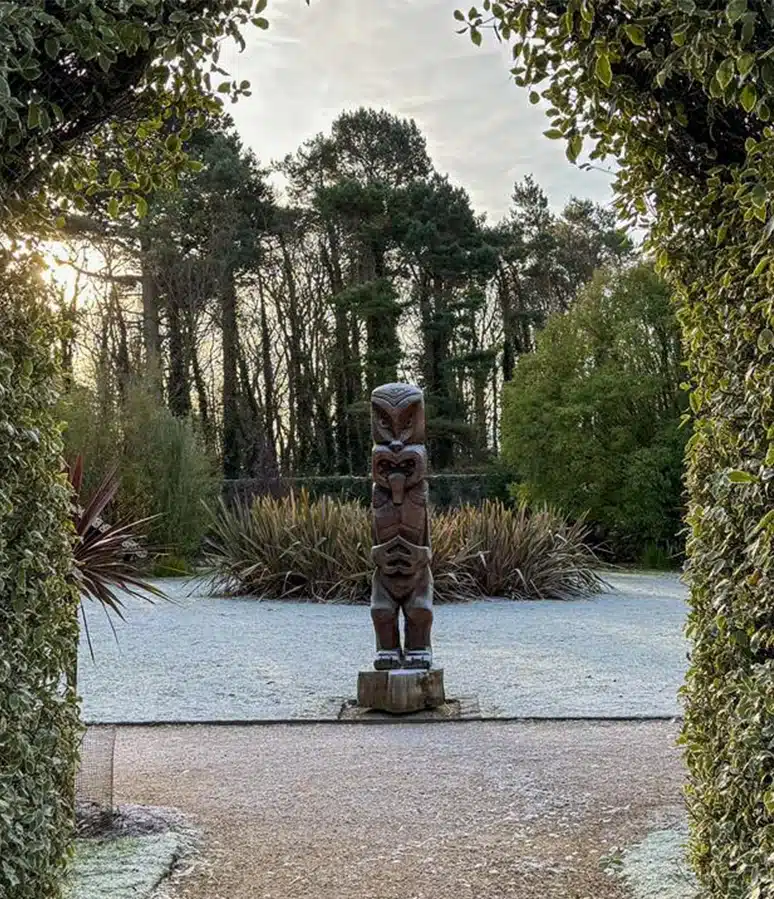 A wooden statue stands on a frost-covered ground framed by an arch of green leafy plants, reminiscent of the enchanting gardens at Malahide Castle. In the background, trees and shrubs are visible under a partly cloudy sky.