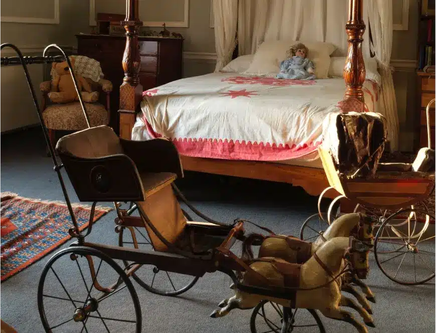 A vintage bedroom reminiscent of grand castle rooms features a canopy bed adorned with a doll, covered by a white and red quilt. Old-fashioned toy horse carriages grace the foreground, while a small woven rug decorates the floor. Completing the scene are a wooden chest and chair.