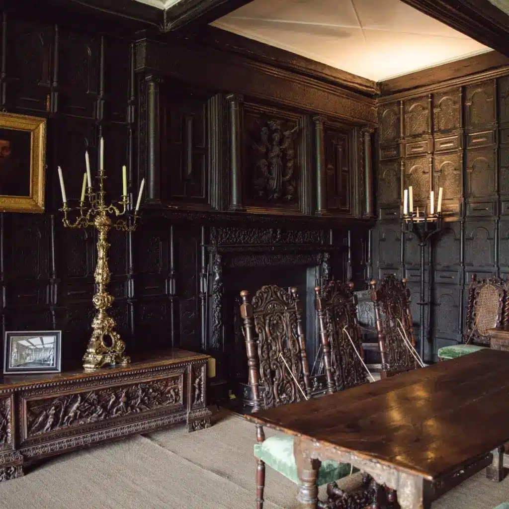 A dimly lit room with ornate wooden paneling, antique chairs, and a large wooden table exudes historical charm. Two golden candelabras by the intricate fireplace highlight details reminiscent of Malahide Castle. A portrait painting completes the scene, perfect for guided tours exploring history's depths.