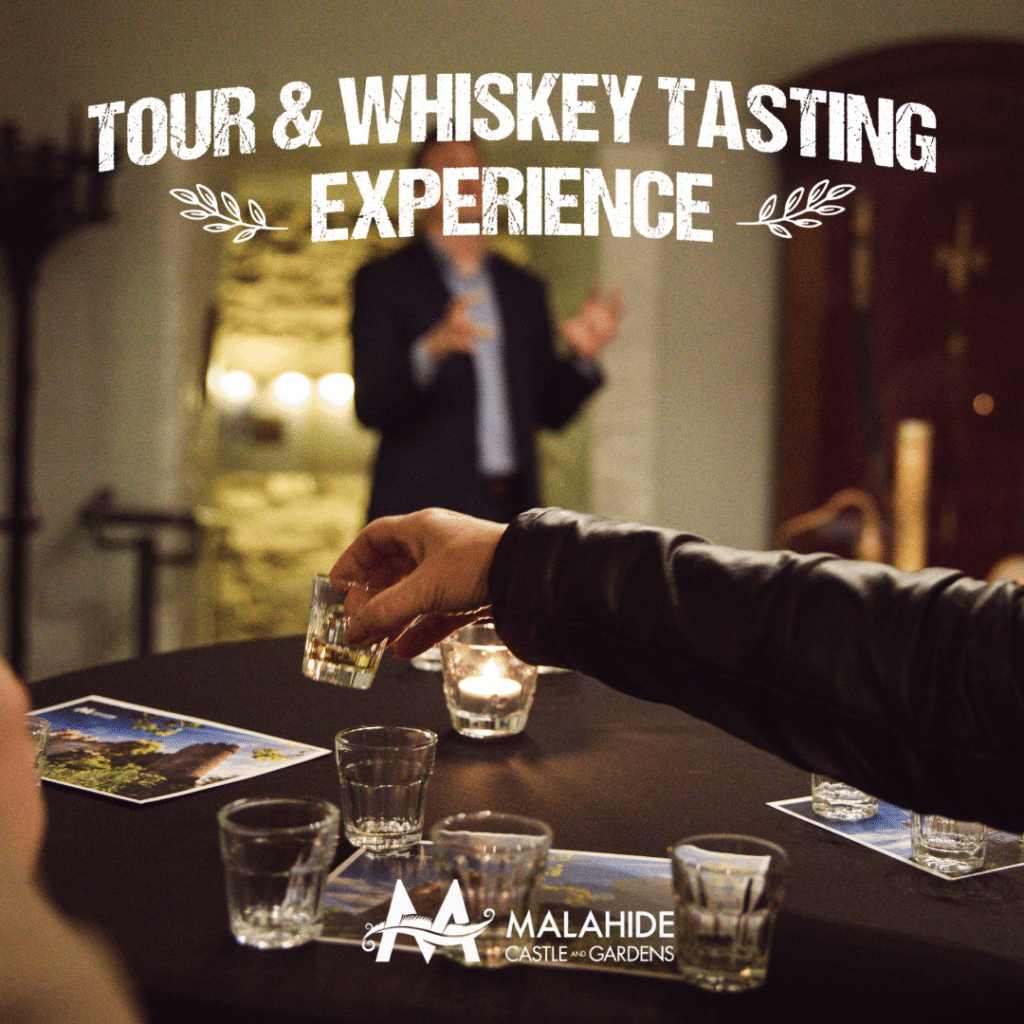 A person holds a glass of whiskey, offering it to another at a tasting event. In the blurred background, someone discusses Malahide Castle & Gardens. A candle flickers beside brochures and a flyer for a thrilling "Tour & Whiskey Tasting Experience" during the Battle of the Bands weekend.