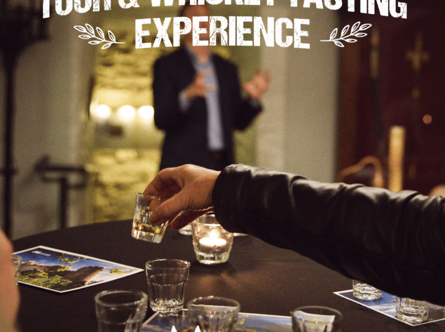 A person holds a glass of whiskey, offering it to another at a tasting event. In the blurred background, someone discusses Malahide Castle & Gardens. A candle flickers beside brochures and a flyer for a thrilling "Tour & Whiskey Tasting Experience" during the Battle of the Bands weekend.