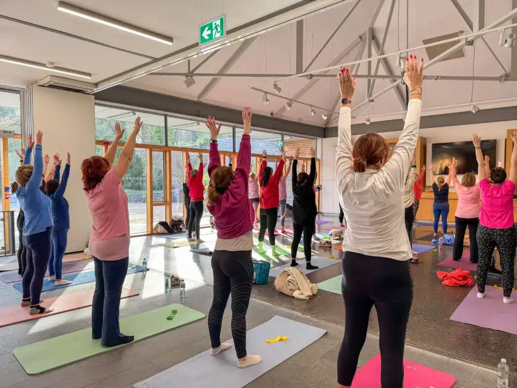A group of people practice yoga indoors, standing on mats with arms raised overhead. Large windows let in sunlight, and yoga props are scattered nearby at this International Women’s Day event. Most participants wear colorful activewear.