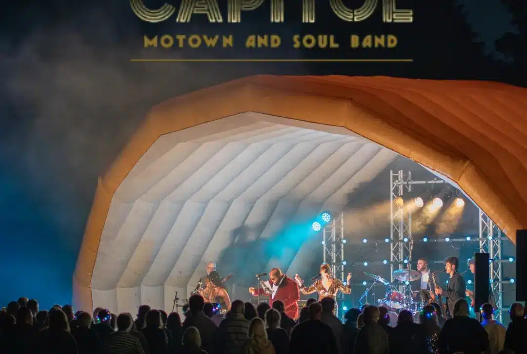 A Motown and soul band enchants the night as they perform under an inflatable shell canopy with "CAPITOL" shining above. While the tour offers soulful tunes, some enjoy a delightful whiskey tasting beneath the blue and purple stage lights.