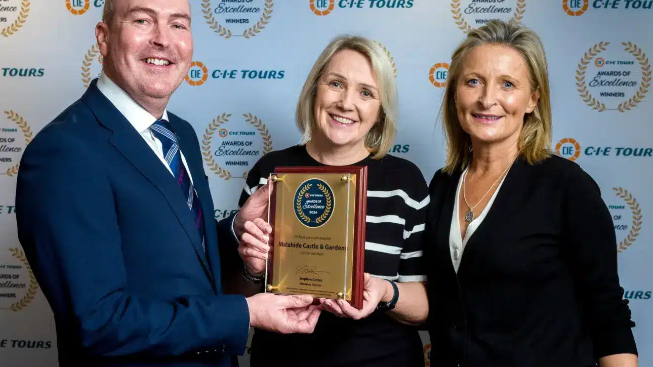 Three people smiling and holding a plaque at an awards event. The man on the left is in a suit and tie, while the two women don formal attire. A backdrop with CIE Tours logos for advertising.