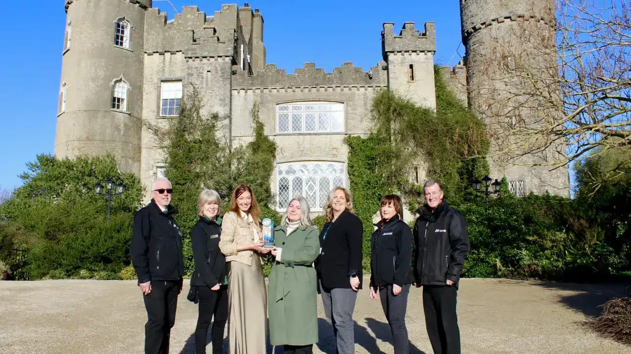 A group of seven people stands in front of a historic, ivy-covered stone castle with turrets, posing together for the camera with an award. The central figure holds an award, all dressed in winter clothing under the clear blue sky, with bare trees visible to the right.