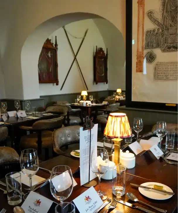 Elegant restaurant interior at Nugents Hall Malahide Castle with set tables, wine glasses, and place cards. Soft lighting from table lamps; walls adorned with crossed swords, shields, and artwork. Cozy and inviting atmosphere.